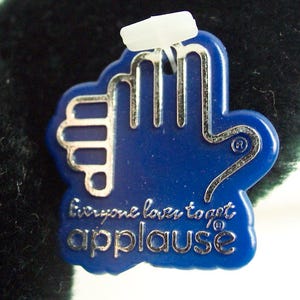 May include: Blue plastic tag with a silver hand outline and the text "Everyone loves to get applause" in silver lettering.