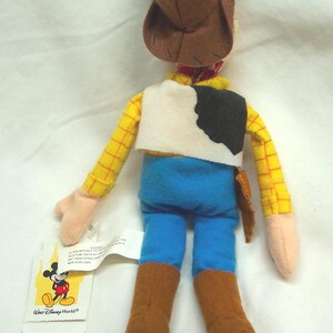 May include: A plush Woody doll from Disney's Toy Story. The doll features a brown cowboy hat, yellow plaid shirt, blue pants, and brown boots. It has a white vest with a black cowhide pattern and a small holster. A Disney World tag is attached.