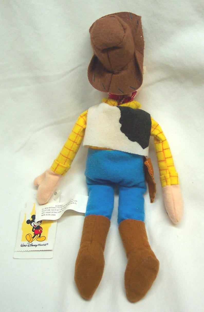 May include: A plush Woody doll from Disney's Toy Story. The doll features a brown cowboy hat, yellow plaid shirt, blue pants, and brown boots. It has a white vest with a black cowhide pattern and a small holster. A Disney World tag is attached.