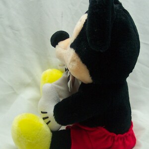 Pu&ograve; includere: Un giocattolo di peluche di Topolino seduto con le gambe incrociate. Topolino indossa pantaloncini rossi e ha la pelliccia nera. Il giocattolo &egrave; alto circa 30 cm.