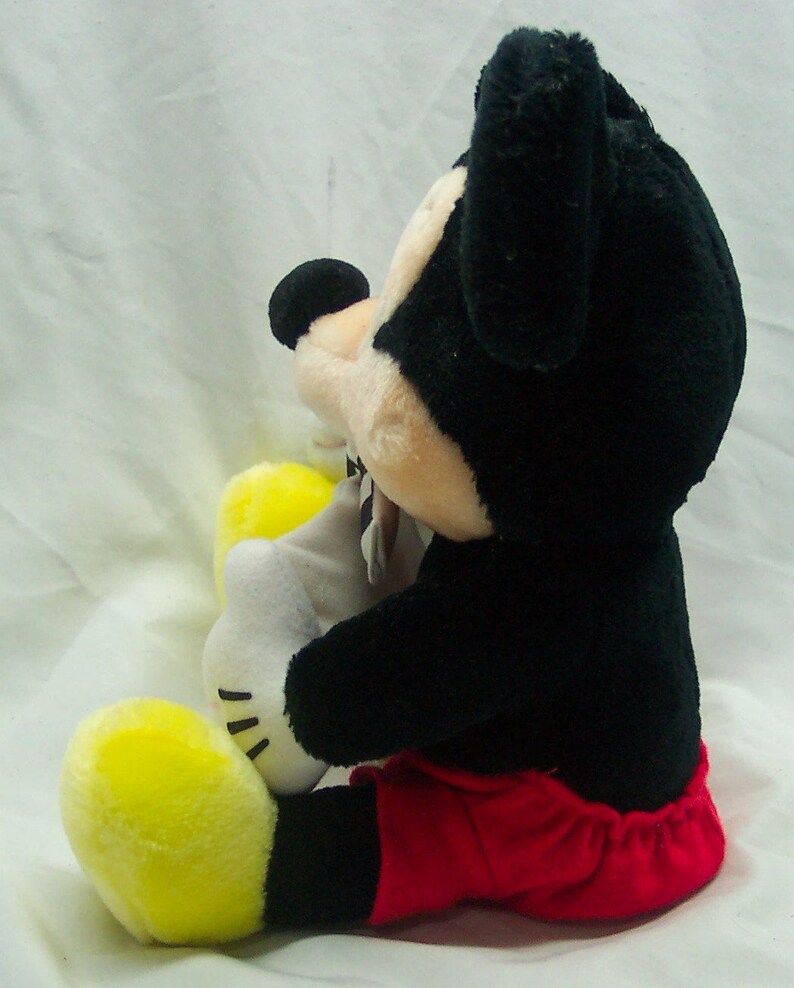 Pu&ograve; includere: Un giocattolo di peluche di Topolino seduto con le gambe incrociate. Topolino indossa pantaloncini rossi e ha la pelliccia nera. Il giocattolo &egrave; alto circa 30 cm.