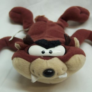 May include: A plush toy of the Tasmanian Devil, a cartoon character. The toy is primarily brown and tan, with large white eyes, a black nose, and a toothy grin. It has outstretched arms and legs.
