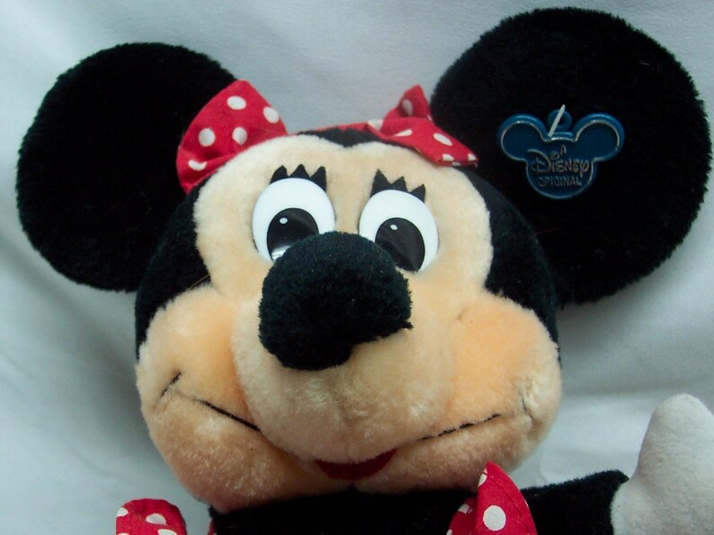 May include: A plush Minnie Mouse toy with a red and white polka dot bow, large black eyes, and a black nose. The toy has a Disney Original tag attached to one ear.