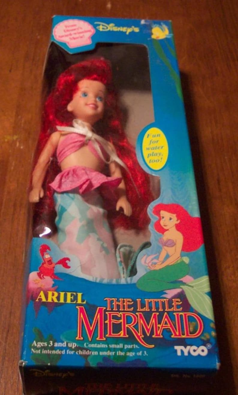 May include: A blue and white box with a picture of Ariel from The Little Mermaid on the front. The box is for a doll that is dressed in a pink and blue mermaid costume. The box says "Ariel The Little Mermaid" and "Fun for water play, too!"