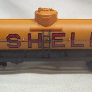 May include: A yellow toy train car with black wheels and a black undercarriage. The car is a tanker car with the word "SHELL" printed in red letters on the side.