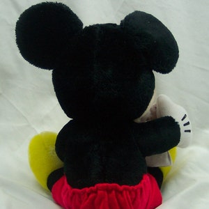 Pu&ograve; includere: Un giocattolo di peluche di Topolino seduto con le gambe incrociate. Il giocattolo &egrave; nero con grandi orecchie nere, scarpe gialle e pantaloncini rossi e bianchi.