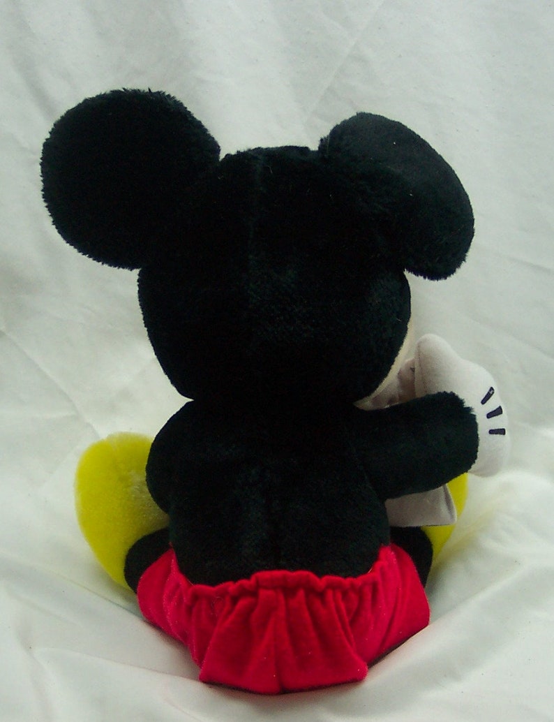 Pu&ograve; includere: Un giocattolo di peluche di Topolino seduto con le gambe incrociate. Il giocattolo &egrave; nero con grandi orecchie nere, scarpe gialle e pantaloncini rossi e bianchi.