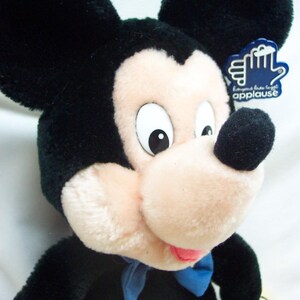 May include: Black and white plush Mickey Mouse toy wearing a blue bow tie and red shorts with a yellow button. The toy has a tag that says "Give us a big hand applause".