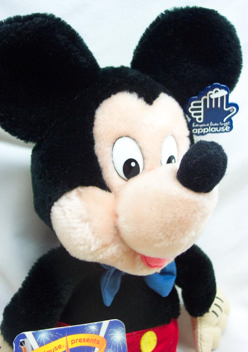 May include: Black and white plush Mickey Mouse toy wearing a blue bow tie and red shorts with a yellow button. The toy has a tag that says "Give us a big hand applause".