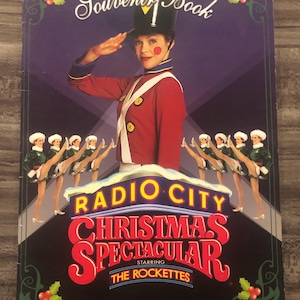 May include: Souvenir book cover for the Radio City Christmas Spectacular, featuring a woman in a red and white uniform saluting. The title is in red and yellow, with the Rockettes performing below.