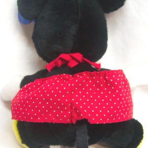 May include: A plush Minnie Mouse doll, viewed from the back, featuring black fur, large round ears, and a red polka-dot skirt. A red bow is at the neck, and a black tail extends downward. The doll has white gloves and yellow shoes.