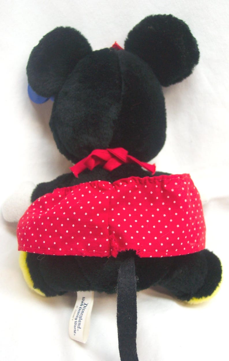 May include: A plush Minnie Mouse doll, viewed from the back, featuring black fur, large round ears, and a red polka-dot skirt. A red bow is at the neck, and a black tail extends downward. The doll has white gloves and yellow shoes.