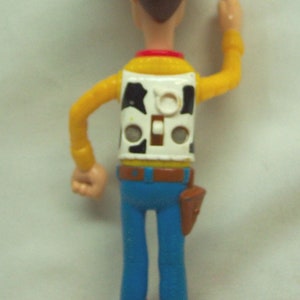 Pu&ograve; includere: Una figurina di plastica di Woody del franchise Toy Story. Woody indossa il suo cappello da cowboy marrone distintivo, una camicia gialla, un gilet bianco con un motivo di mucca bianco e nero, jeans blu e stivali da cowboy marroni. Sta alzando il braccio destro in aria, come se stesse salutando.
