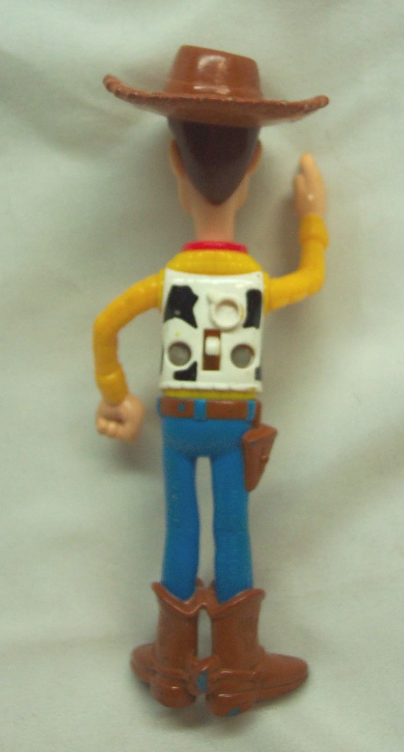 Pu&ograve; includere: Una figurina di plastica di Woody del franchise Toy Story. Woody indossa il suo cappello da cowboy marrone distintivo, una camicia gialla, un gilet bianco con un motivo di mucca bianco e nero, jeans blu e stivali da cowboy marroni. Sta alzando il braccio destro in aria, come se stesse salutando.