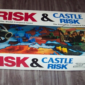 May include: A rectangular board game box for Risk and Castle Risk by Parker Brothers. The white box features red and blue text, a graphic of a black cannon, a map of Europe and North Africa, and yellow star-shaped game pieces on the lid.