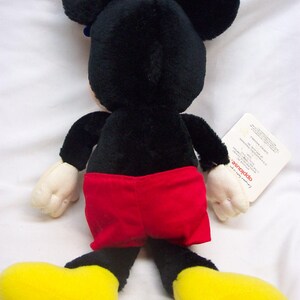 May include: A black and red plush Mickey Mouse toy with yellow shoes. The toy is sitting on a white surface and has a tag attached to its right leg.