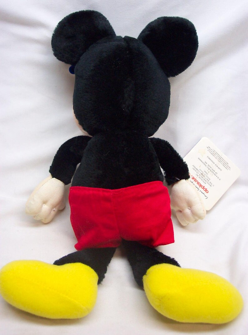 May include: A black and red plush Mickey Mouse toy with yellow shoes. The toy is sitting on a white surface and has a tag attached to its right leg.