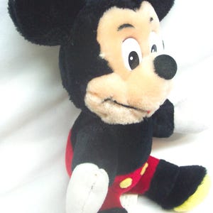 Pode incluir: Uma boneca de pel&uacute;cia do Mickey Mouse, com pelo preto, orelhas grandes e redondas e um rosto bege com um nariz preto. A boneca usa cal&ccedil;&otilde;es vermelhos com bot&otilde;es amarelos e sapatos com solas amarelas. O brinquedo est&aacute; sobre um fundo branco.