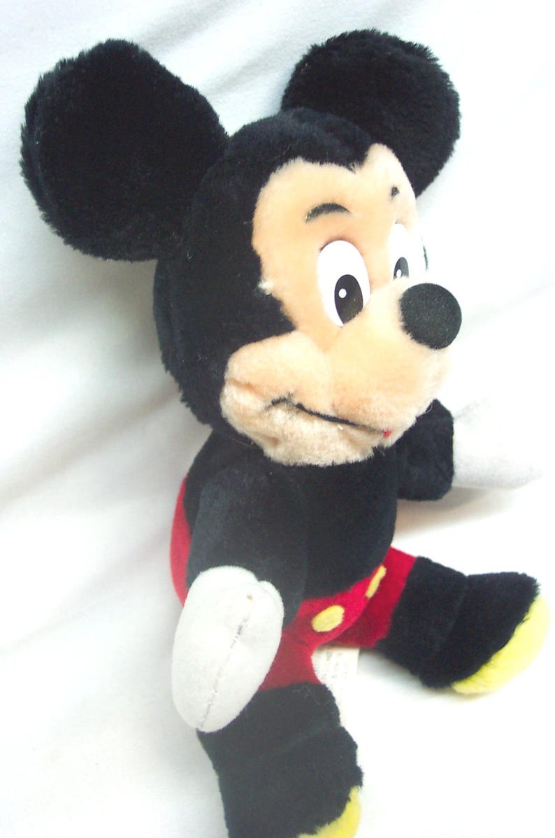 Pode incluir: Uma boneca de pel&uacute;cia do Mickey Mouse, com pelo preto, orelhas grandes e redondas e um rosto bege com um nariz preto. A boneca usa cal&ccedil;&otilde;es vermelhos com bot&otilde;es amarelos e sapatos com solas amarelas. O brinquedo est&aacute; sobre um fundo branco.