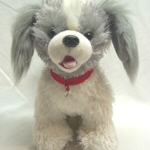 Può includere: Un peluche a forma di cane con pelo grigio arruffato sulla testa e sulle orecchie, una macchia bianca sulla fronte e pelo color crema sul corpo. Il giocattolo presenta un naso nero, una bocca aperta con una lingua rosa e un collare rosso brillante con un piccolo anello di metallo.