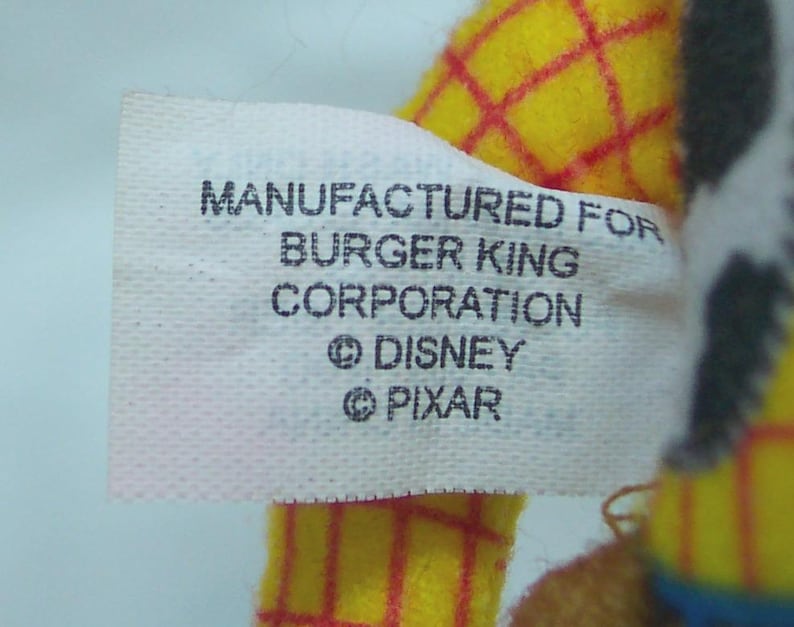 Puede incluir: Primer plano de una etiqueta blanca con texto negro que dice "MANUFACTURED FOR BURGER KING CORPORATION &copy; DISNEY &copy; PIXAR". La etiqueta est&aacute; adherida a un juguete de peluche amarillo con l&iacute;neas de cuadr&iacute;cula rojas.