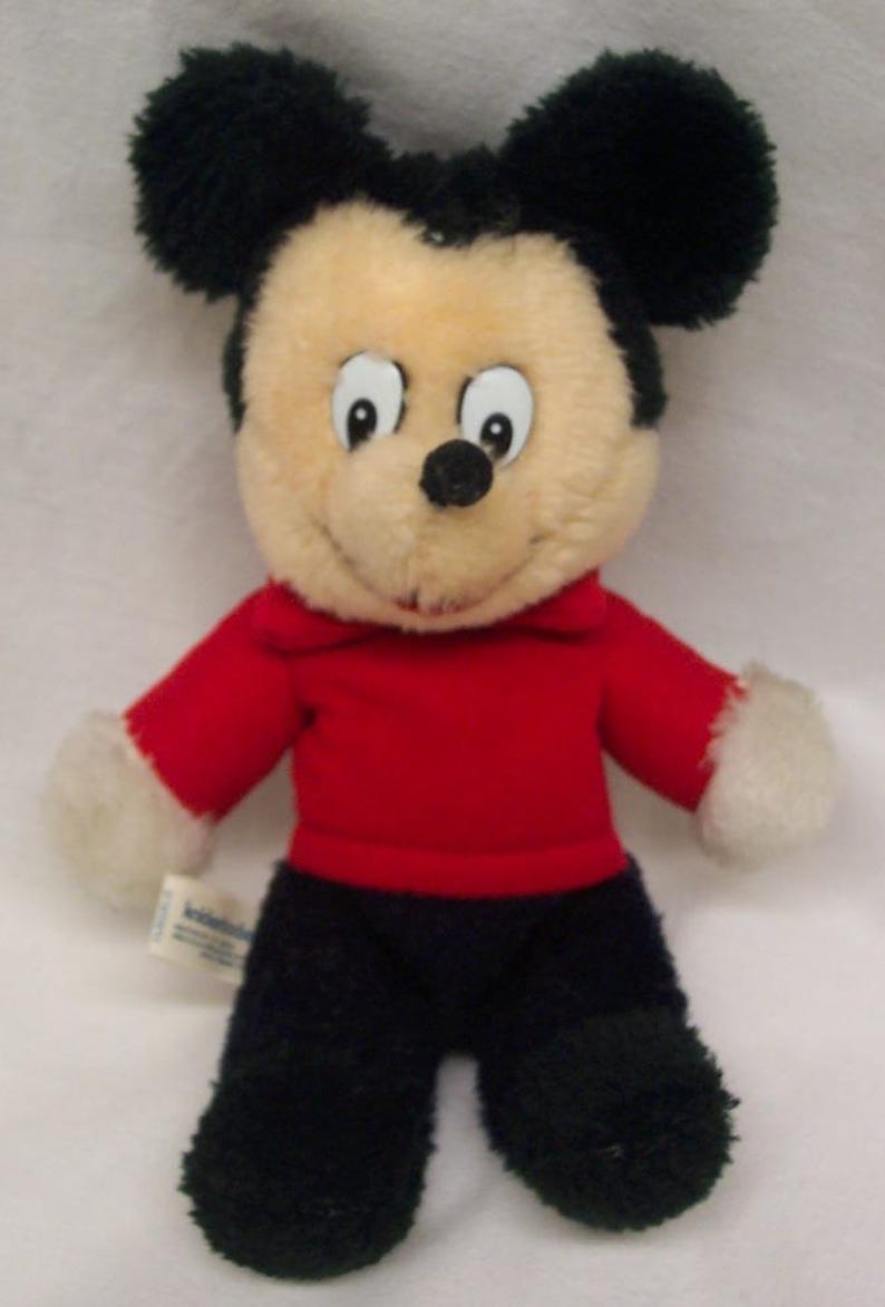 May include: A plush Mickey Mouse toy wearing a red shirt and black pants. The toy has black fur, a black nose, and white fur on its hands and feet. The toy has a tag on its left leg that says 'Knickerbocker'.