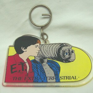 May include: A clear acrylic keychain featuring a graphic of a young person and the character E.T. against a yellow background. The text E.T. THE EXTRA-TERRESTRIAL is printed in black. The keychain includes a silver-toned metal split ring for keys.