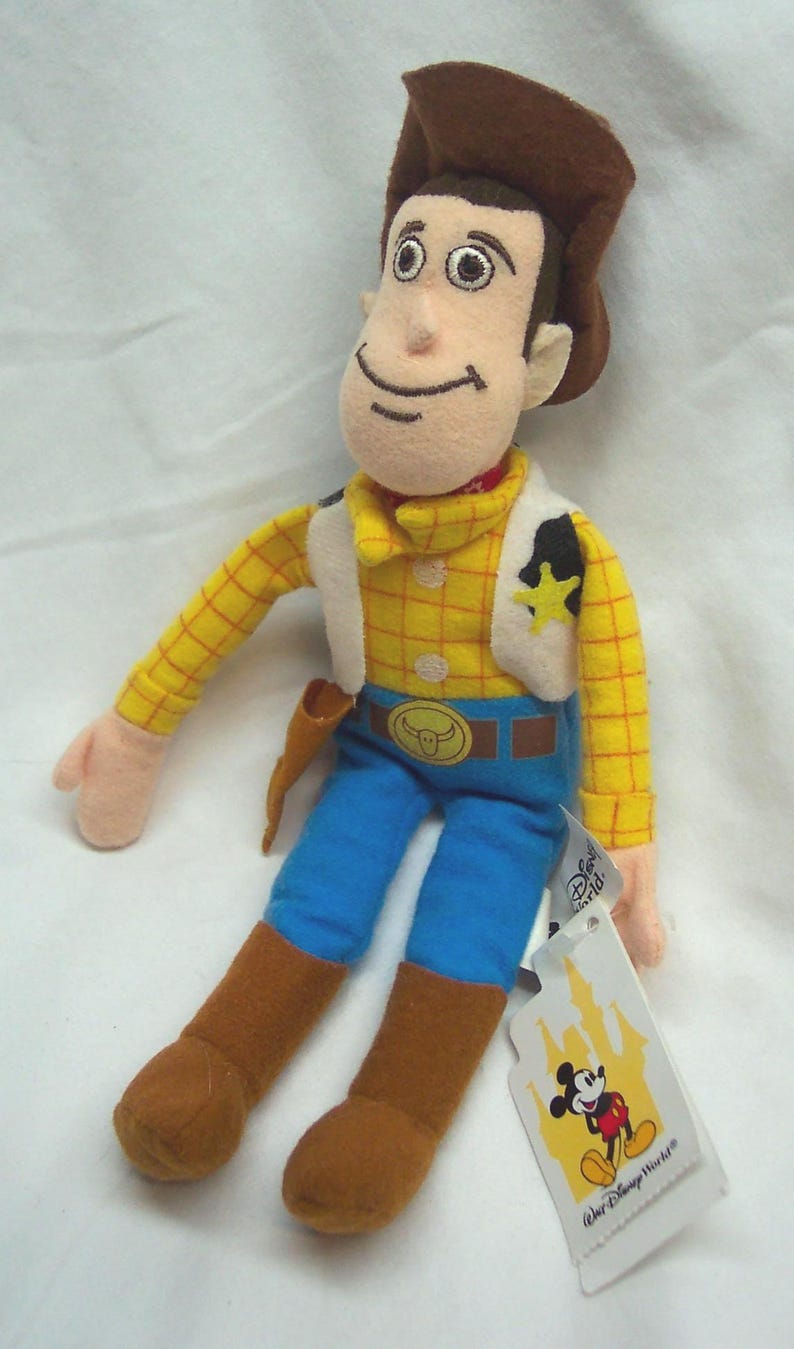 May include: A plush Woody doll from Disney's Toy Story. The doll is wearing a brown cowboy hat, yellow plaid shirt, blue pants, and brown boots. It has a white vest, a sheriff's badge, and a belt buckle. A Disney World tag is attached.