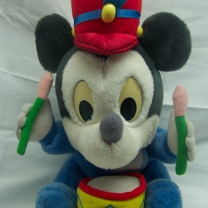 May include: A plush Mickey Mouse toy dressed as a drummer, wearing a red and blue hat with a feather, holding two drumsticks and sitting on a yellow and red drum.