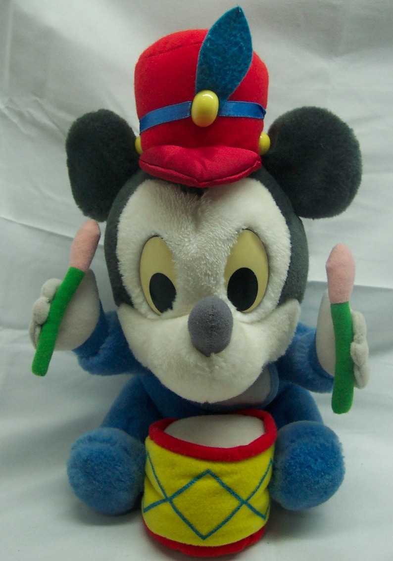 May include: A plush Mickey Mouse toy dressed as a drummer, wearing a red and blue hat with a feather, holding two drumsticks and sitting on a yellow and red drum.