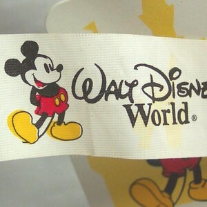 May include: A white tag featuring the Walt Disney World logo and a cartoon image of Mickey Mouse. Mickey Mouse is depicted in his classic outfit, with red shorts, yellow shoes, and black gloves. The tag is partially visible against a blue background.