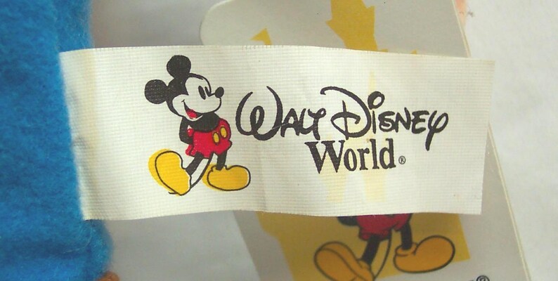 May include: A white tag featuring the Walt Disney World logo and a cartoon image of Mickey Mouse. Mickey Mouse is depicted in his classic outfit, with red shorts, yellow shoes, and black gloves. The tag is partially visible against a blue background.