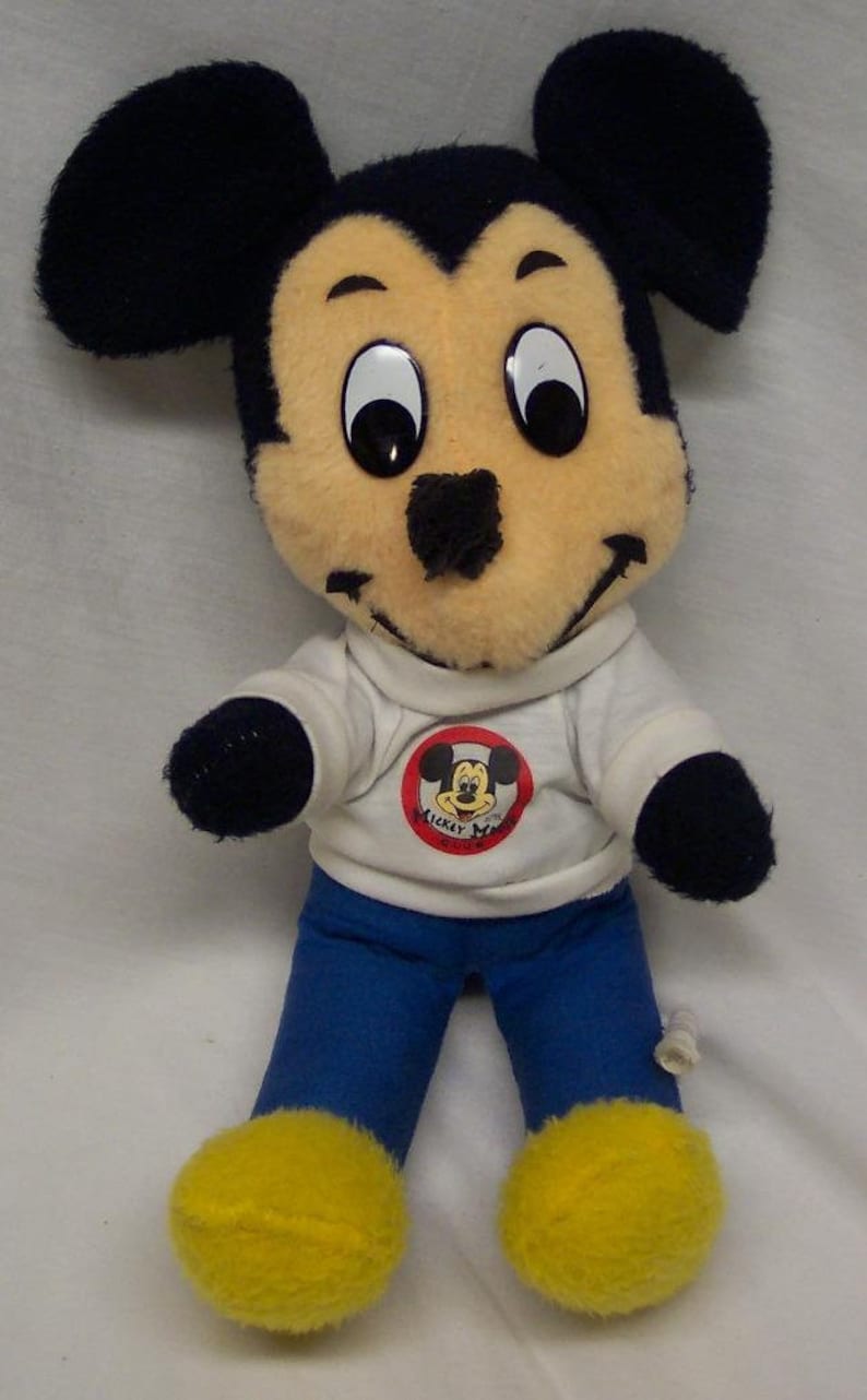 K&ouml;nnte beinhalten: Ein Pl&uuml;sch-Mickey-Mouse-Spielzeug, das ein wei&szlig;es Hemd mit einem roten Mickey-Mouse-Logo und blaue Hosen tr&auml;gt. Das Spielzeug hat gro&szlig;e schwarze Ohren, schwarze Augen und gelbe Schuhe.