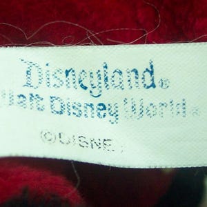 Pode incluir: Close-up de uma etiqueta branca com as palavras "Disneyland" e "Walt Disney World" impressas em azul. A etiqueta est&aacute; em um fundo vermelho e preto, com o s&iacute;mbolo de copyright da Disney.