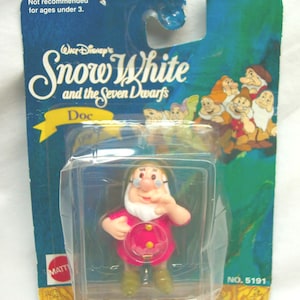 May include: A vintage Mattel action figure of Doc from Disney's Snow White and the Seven Dwarfs, still in its original packaging. The figure is wearing a pink outfit and green shoes. The packaging is blue and features the film's title and characters.