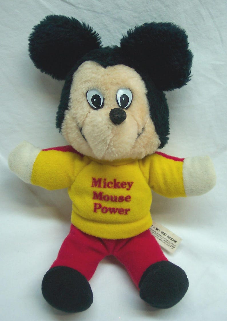 May include: A vintage Mickey Mouse plush toy. The plush has a cream-colored face, black ears, and a yellow shirt with the text "Mickey Mouse Power." It is wearing red pants and black shoes. The toy is a classic Disney collectible.
