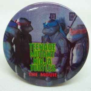 May include: A round button featuring the Teenage Mutant Ninja Turtles from the movie. The button shows the turtles with their colored masks and the movie title in green and red text.