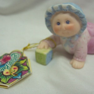 May include: A crawling Cabbage Patch Kids figurine, wearing a pink outfit and a blue bonnet, reaching for a green and blue block. A colorful tag with the Cabbage Patch Kids logo is attached.
