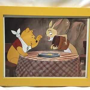 May include: A framed print featuring Winnie the Pooh and Rabbit. Pooh, wearing a bib, sits at a checkered table, while Rabbit pours honey from a jar labelled "Hunny." The frame is yellow.