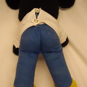K&ouml;nnte beinhalten: Ein schwarzes und wei&szlig;es Pl&uuml;schmaus-Spielzeug, das ein wei&szlig;es Hemd und eine blaue Jeans tr&auml;gt. Die Maus hat gro&szlig;e Ohren und gelbe F&uuml;&szlig;e.