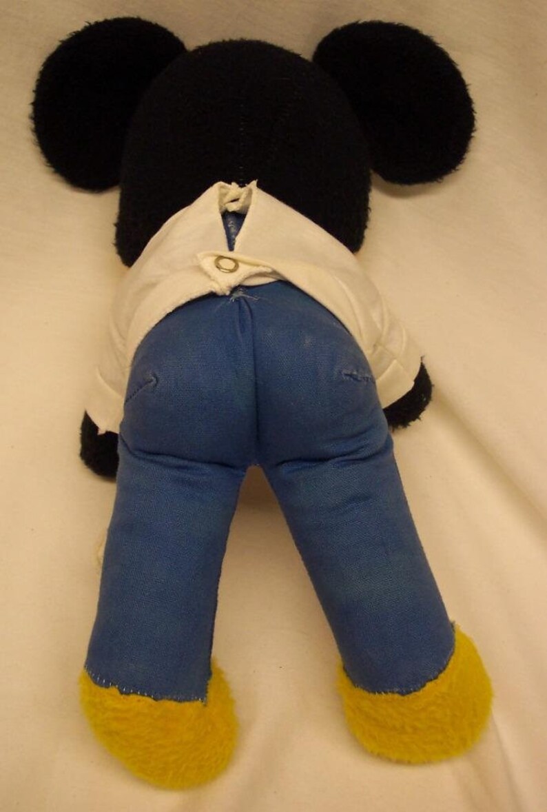 K&ouml;nnte beinhalten: Ein schwarzes und wei&szlig;es Pl&uuml;schmaus-Spielzeug, das ein wei&szlig;es Hemd und eine blaue Jeans tr&auml;gt. Die Maus hat gro&szlig;e Ohren und gelbe F&uuml;&szlig;e.