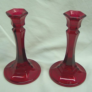 May include: Two ruby red glass candle holders. Each has a hexagonal top, a slender, curved stem, and a round base. The candle holders are made of a transparent, glossy glass material.
