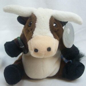 May include: A plush toy cow with brown and white fur, black hooves, and large white horns. The cow has a light pink muzzle and black eyes. A small Coca-Cola bottle is attached to one leg. A tag is attached to the cow's ear.