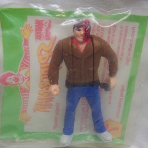 May include: A brown plastic action figure of a man wearing a brown jacket, blue jeans, and a red and black mask. The figure is holding a small black object in his right hand. The figure is packaged in a clear plastic bag with a green and yellow background. The bag has the words "Comics" and "Man" printed on it.