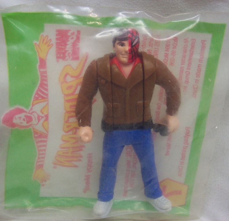 Pu&ograve; includere: Una action figure di plastica marrone di un uomo che indossa una giacca marrone, jeans blu e una maschera rossa e nera. La figura tiene un piccolo oggetto nero nella mano destra. La figura &egrave; confezionata in un sacchetto di plastica trasparente con uno sfondo verde e giallo. Il sacchetto ha le parole "Comics" e "Man" stampate su di esso.