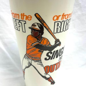 May include: Cream-colored baseball cup featuring a baseball player in an orange uniform with black accents, holding a bat. Text on the cup reads "From the Left or from Right, Sing Out!" A vintage sports collectible.