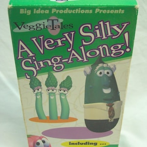 May include: A VHS tape for "VeggieTales: A Very Silly Sing-Along!" featuring cartoon vegetables. The box is green and white with the title in large purple letters. The cover includes illustrations of asparagus, a cucumber, and a tomato.
