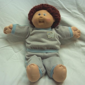 Peut inclure: Une poupée Cabbage Patch Kid aux cheveux bruns portant un pyjama gris et bleu avec un patch de chat. La poupée a un teint rosé et des yeux bleus.