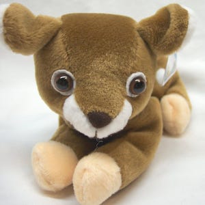 May include: A plush toy shaped like a fawn with soft brown fur, large dark brown eyes, and white markings around the muzzle and ears. The stuffed animal is in a lying position with light tan paws and a small dark brown nose, featuring a tag.