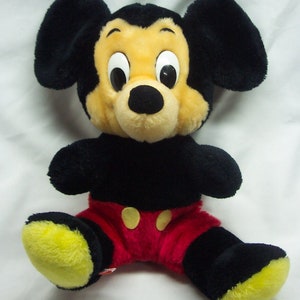 May include: A plush black and yellow Mickey Mouse toy wearing red shorts with yellow buttons. The toy has large black ears and a black nose.